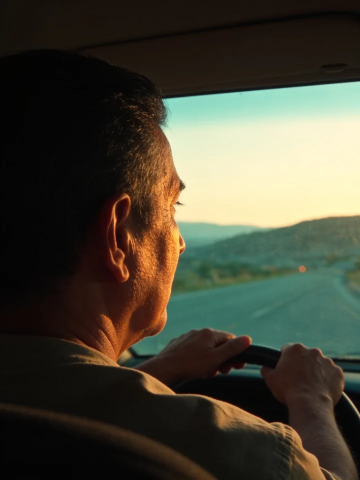 Un hombre conduce de vuelta a casa al atardecer; el sol bajo entra por el parabrisas y la carretera se abre hacia unas colinas.